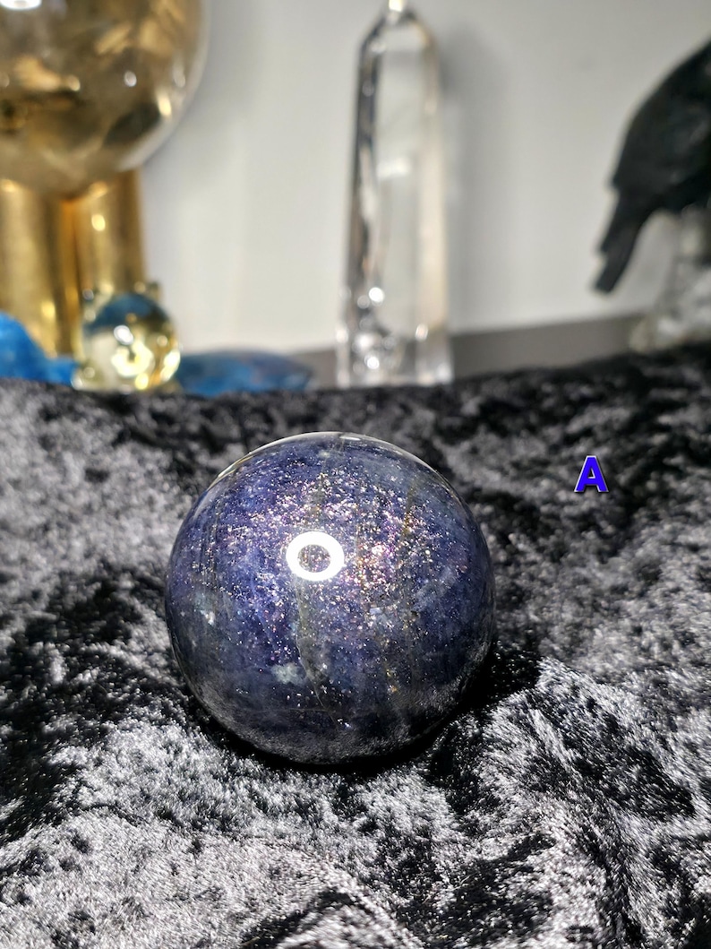 May include: A dark blue crystal sphere with shimmering inclusions. The sphere is set against a black velvet surface. Other objects are visible in the background, including a gold-colored decorative item and a clear glass bottle.