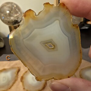 May include: A natural agate slice, displaying a cream and brown colour palette. The stone has a unique, irregular edge and a central, eye-like pattern. The agate is held by a person's fingers, with other slices visible in the background.