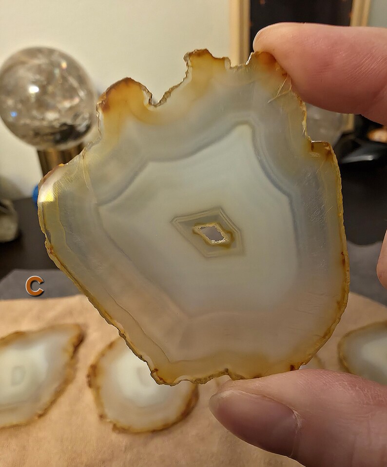 May include: A natural agate slice, displaying a cream and brown colour palette. The stone has a unique, irregular edge and a central, eye-like pattern. The agate is held by a person's fingers, with other slices visible in the background.