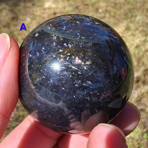 May include: A dark, spherical gemstone with a polished surface. The sphere has a deep blue and black color with shimmering, reflective inclusions. The gem is held in a hand, with the background out of focus.