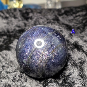 May include: A dark blue crystal ball with a shimmering, speckled surface. The sphere has a polished appearance and is set against a dark, textured background. The letter "A" is visible in the background.