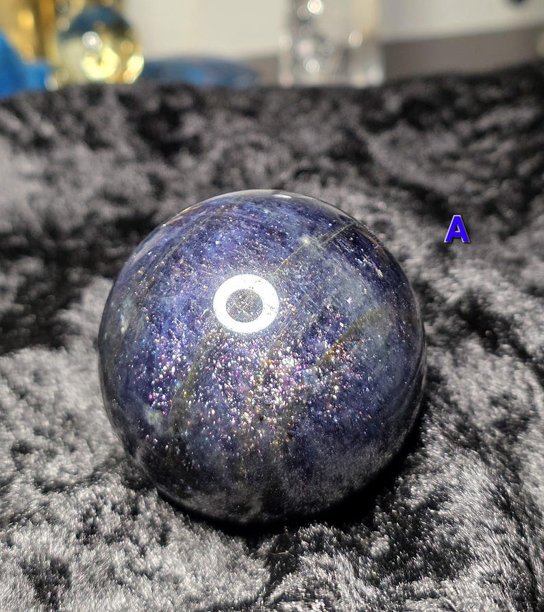 May include: A dark blue crystal ball with a shimmering, speckled surface. The sphere has a polished appearance and is set against a dark, textured background. The letter "A" is visible in the background.