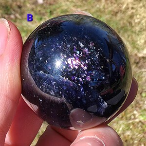 May include: A dark, polished sphere with a reflective surface, displaying a galaxy of purple and iridescent flecks. The letter "B" is visible in the upper left corner. The sphere is held in a hand, with a blurred background of green grass.