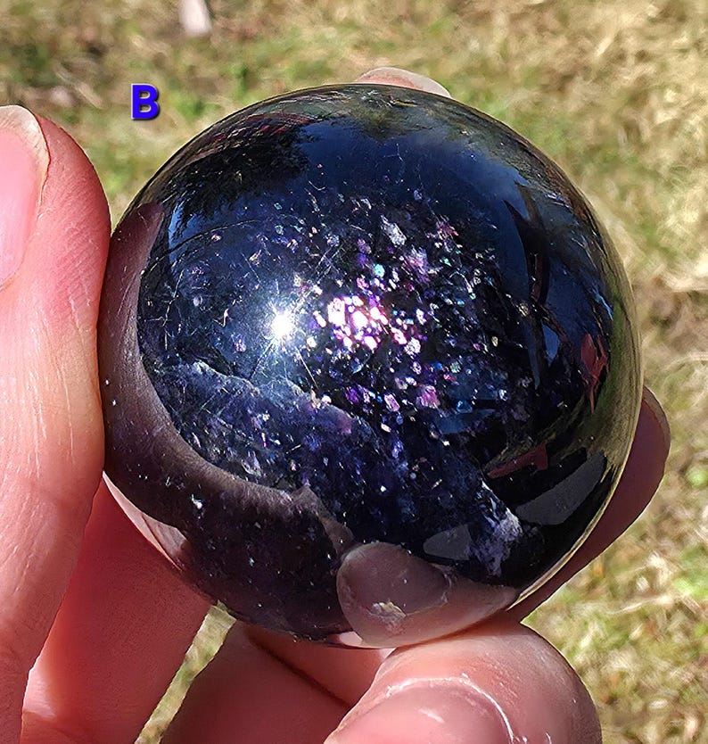 May include: A dark, polished sphere with a reflective surface, displaying a galaxy of purple and iridescent flecks. The letter "B" is visible in the upper left corner. The sphere is held in a hand, with a blurred background of green grass.