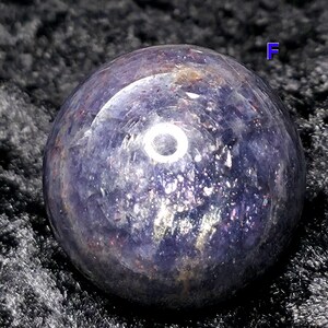 May include: A polished, spherical gemstone with a deep purple hue, speckled with iridescent flecks. The orb reflects light, creating a bright circular highlight. The sphere is set against a dark, textured background.