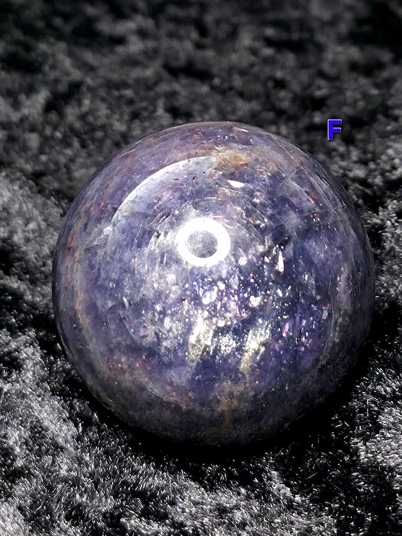 May include: A polished, spherical gemstone with a deep purple hue, speckled with iridescent flecks. The orb reflects light, creating a bright circular highlight. The sphere is set against a dark, textured background.