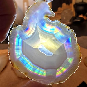May include: A translucent, polished agate slice with a rainbow iridescence. The stone displays concentric bands of white, gray, and brown, with vibrant colors refracting light. The agate is held between fingers, showcasing its natural beauty and unique patterns.