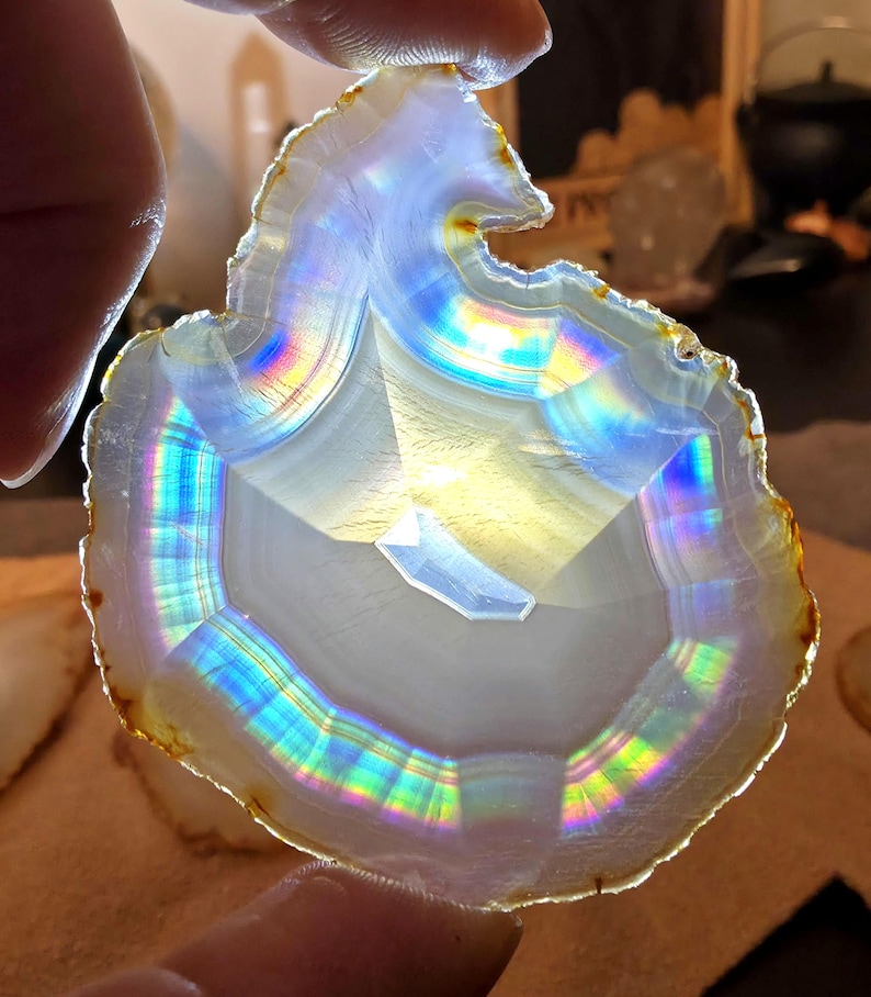 May include: A polished agate slice, displaying a spectrum of colours due to iridescence. The stone features concentric bands of white, grey, and brown. The agate is held between fingers, highlighting its natural patterns and the play of light across its surface.