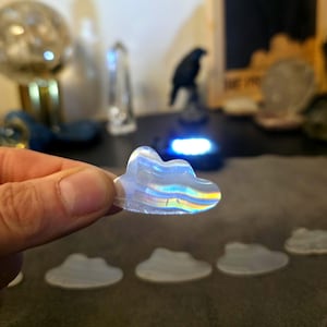 May include: A clear, cloud-shaped crystal with iridescent rainbow hues is held in a hand. Several similar cloud-shaped crystals are visible in the foreground. The background is blurred, with other crystals and a decorative raven statue.