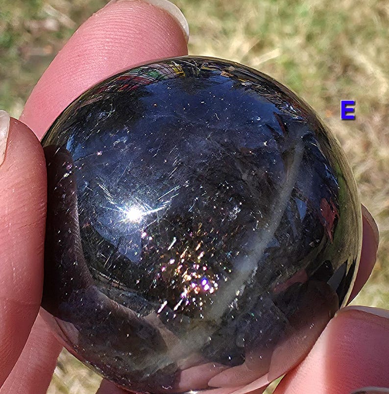 May include: A polished, dark sphere with iridescent flecks of color. The sphere has a reflective surface, with hints of purple, gold, and blue. The sphere is held between two fingers.