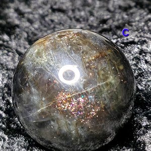 May include: A polished, spherical gemstone with a dark, iridescent surface. The sphere has a mix of colors, including gold, silver, and hints of purple. The gemstone is set against a dark, textured background.