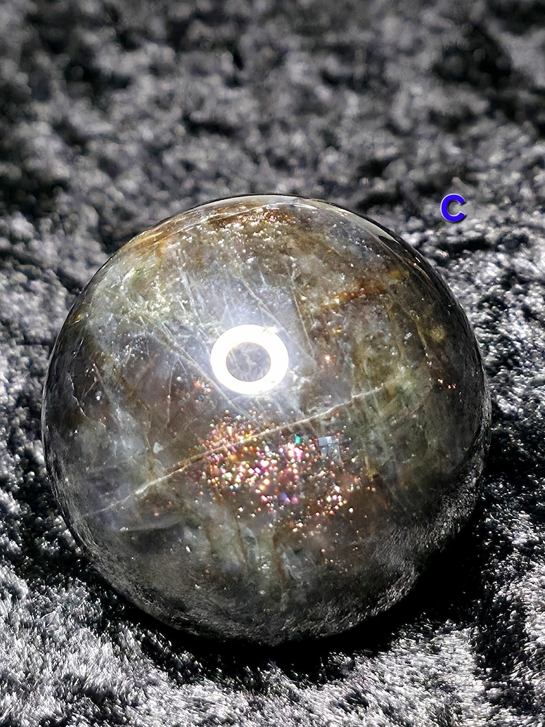 May include: A polished, spherical gemstone with a dark, iridescent surface. The sphere has a mix of colors, including gold, silver, and hints of purple. The gemstone is set against a dark, textured background.