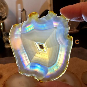 May include: A polished agate slice with a natural, uneven edge. The stone displays concentric bands of white, grey, and iridescent colours, with a central geometric pattern. The edge has a golden-brown hue.