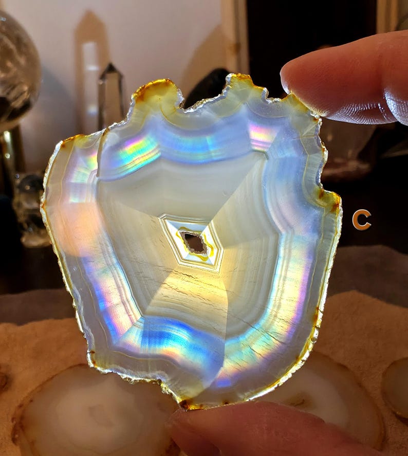 May include: A polished agate slice with a natural, uneven edge. The stone displays concentric bands of white, grey, and iridescent colours, with a central geometric pattern. The edge has a golden-brown hue.