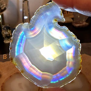May include: A translucent agate slice with a white core and a rainbow-hued border. The agate is held by a finger, highlighting its natural patterns and colours. The slice has a polished surface and a rough, natural edge. The agate is a decorative item.