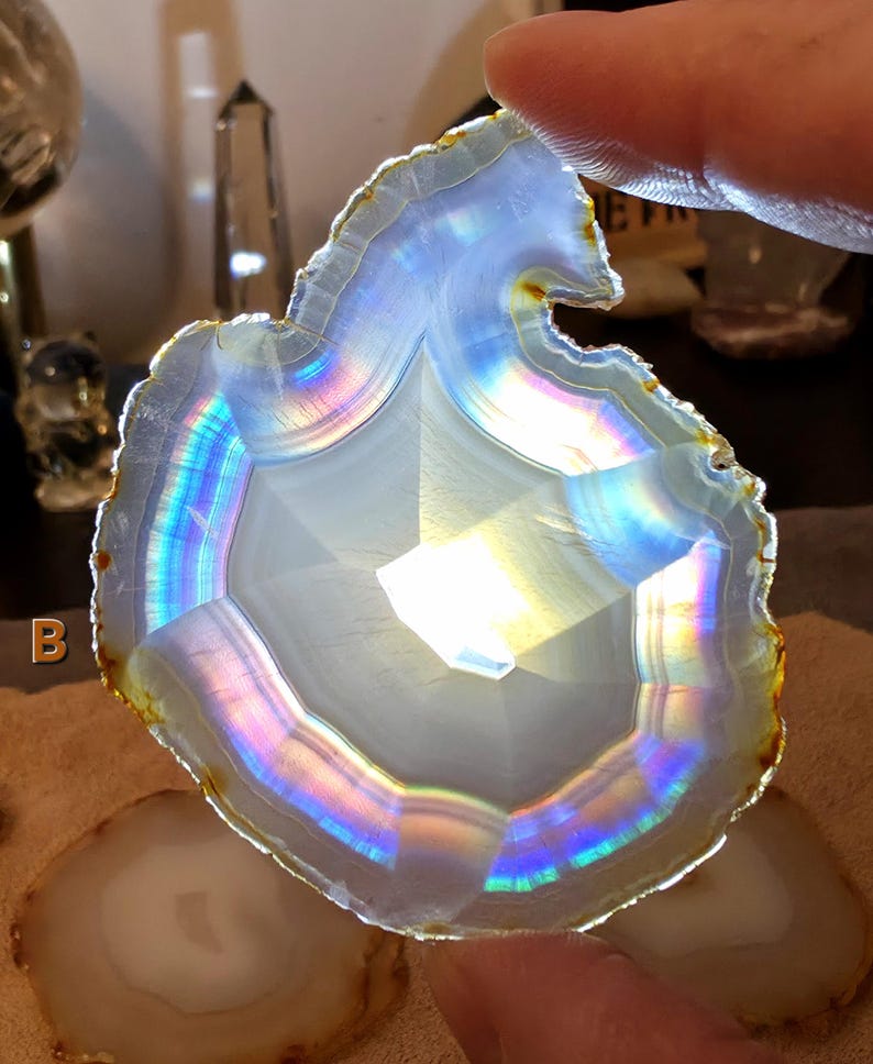 May include: A translucent agate slice with a white core and a rainbow-hued border. The agate is held by a finger, highlighting its natural patterns and colours. The slice has a polished surface and a rough, natural edge. The agate is a decorative item.