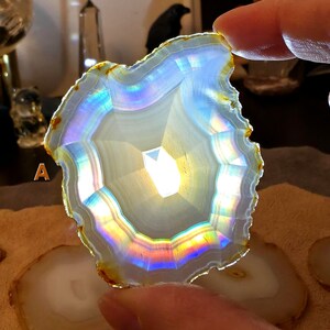 May include: A translucent, polished agate slice with a white centre and iridescent rainbow hues. The outer edge is a golden-brown colour. The agate is held up, with other slices visible in the background. The overall theme is crystals and gemstones.