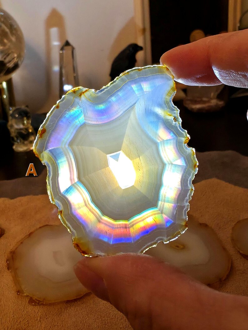 May include: A translucent, polished agate slice with a white centre and iridescent rainbow hues. The outer edge is a golden-brown colour. The agate is held up, with other slices visible in the background. The overall theme is crystals and gemstones.