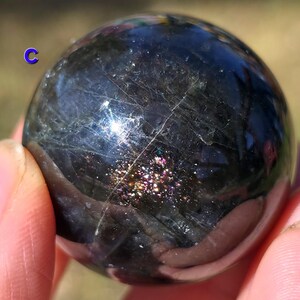 May include: A dark, polished sphere with iridescent inclusions and subtle veining. The sphere is held in a hand, showcasing its smooth, reflective surface. The overall color is a deep, charcoal gray with flashes of color.