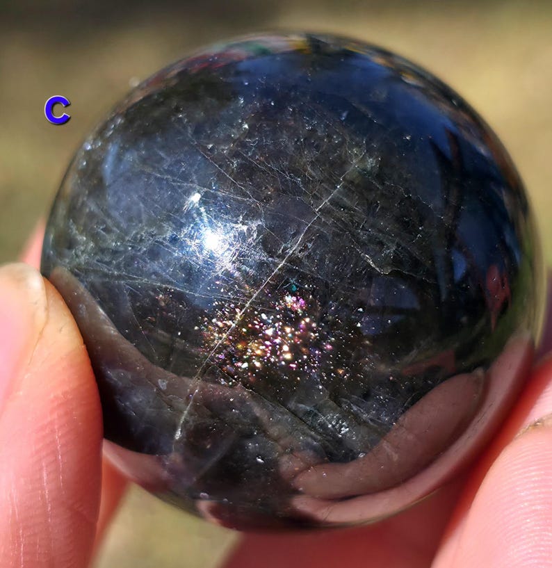 May include: A dark, polished sphere with iridescent inclusions and subtle veining. The sphere is held in a hand, showcasing its smooth, reflective surface. The overall color is a deep, charcoal gray with flashes of color.