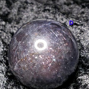 May include: A dark, iridescent sphere with a polished surface. The sphere has a mix of dark purple and silver tones, with sparkling inclusions throughout. The object is set against a dark, textured background.
