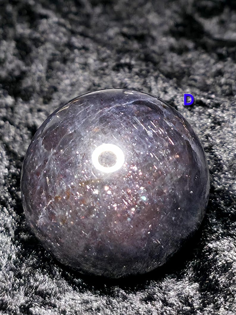May include: A dark, iridescent sphere with a polished surface. The sphere has a mix of dark purple and silver tones, with sparkling inclusions throughout. The object is set against a dark, textured background.