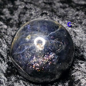 May include: A dark, iridescent crystal ball with a polished surface. The sphere has a deep blue hue with shimmering, colorful inclusions. A bright, circular reflection is visible on the surface, adding a focal point to the crystal.