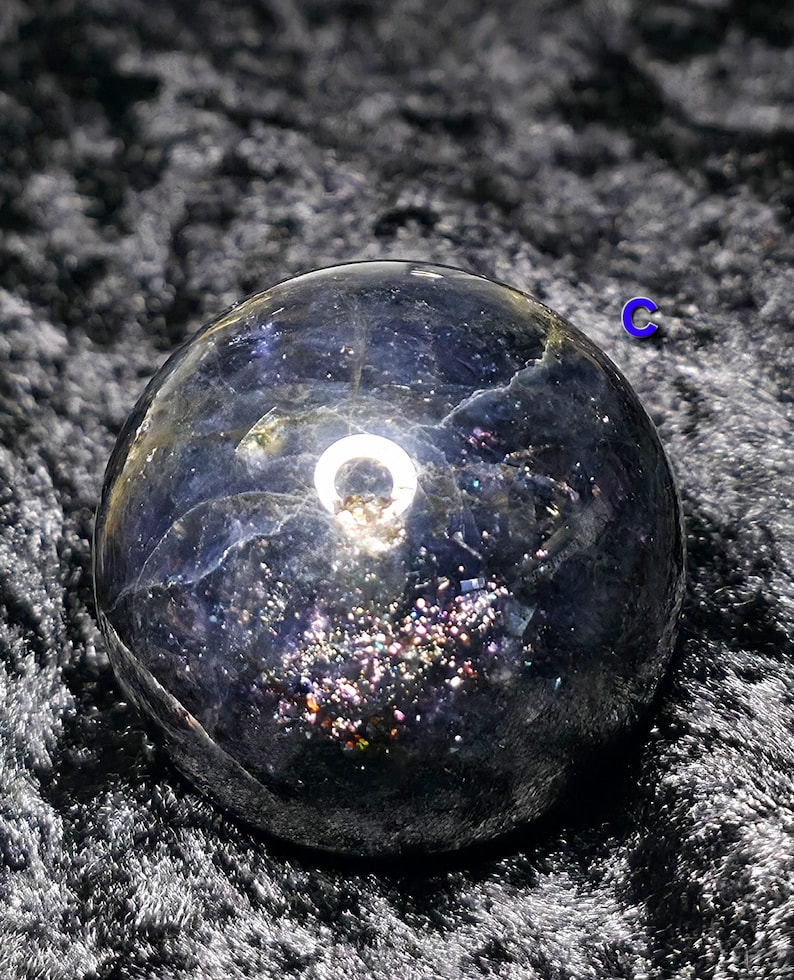 May include: A dark, iridescent crystal ball with a polished surface. The sphere has a deep blue hue with shimmering, colorful inclusions. A bright, circular reflection is visible on the surface, adding a focal point to the crystal.