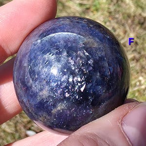 May include: A polished, spherical gemstone with a deep blue and purple color scheme. The surface of the sphere has a reflective, crystalline appearance, with areas of lighter and darker hues. The sphere is held in a hand, with the background out of focus.