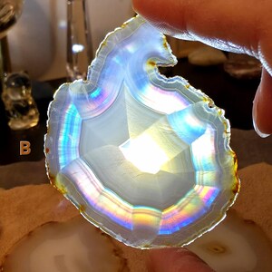 May include: A polished agate slice, displaying a spectrum of colours due to iridescence. The stone has a natural, uneven edge and a central crystalline pattern. The agate is held by a finger, highlighting its light-reflecting qualities. Other agate slices are visible in the background.