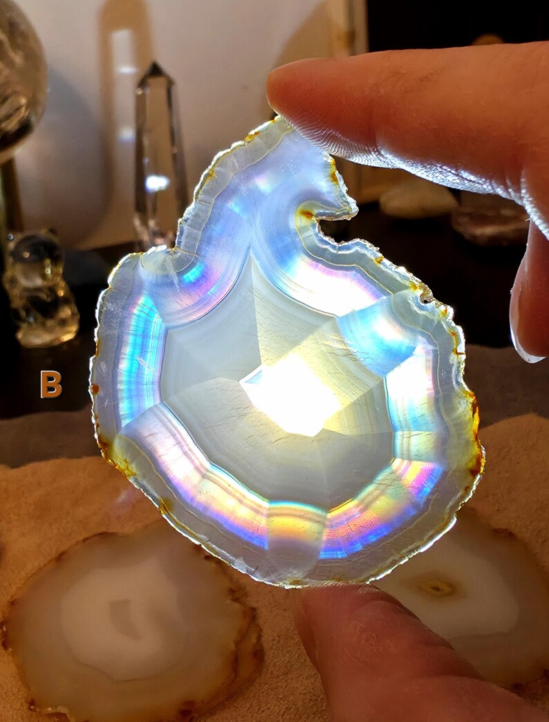 May include: A polished agate slice, displaying a spectrum of colours due to iridescence. The stone has a natural, uneven edge and a central crystalline pattern. The agate is held by a finger, highlighting its light-reflecting qualities. Other agate slices are visible in the background.