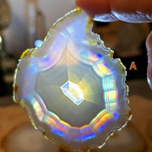 May include: A polished agate slice, showing a central crystalline structure. The stone has concentric bands of white, grey, and yellow, with iridescent rainbow colours along the edges. The agate is held by a person's fingers.