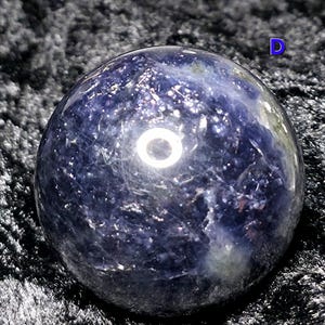 May include: A polished, spherical gemstone with a deep blue hue, speckled with lighter shades and reflective inclusions. The orb appears to be resting on a dark, textured surface, creating a contrast that highlights its form.