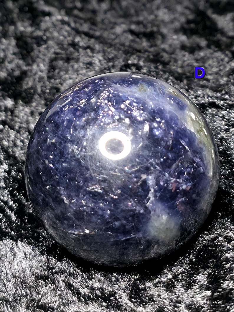 May include: A polished, spherical gemstone with a deep blue hue, speckled with lighter shades and reflective inclusions. The orb appears to be resting on a dark, textured surface, creating a contrast that highlights its form.