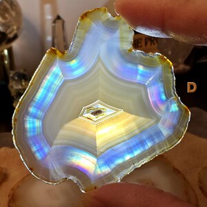 May include: A polished agate slice with a layered, banded pattern. The stone features iridescent bands of blue, yellow, and white, with a central geometric design. The outer edge has a golden-brown hue, and the overall shape is irregular.