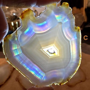May include: A translucent agate slice with a natural, irregular edge. The stone features concentric bands of white and grey, with iridescent rainbow hues reflecting light. The outer edge has a golden-brown colour. The letter "C" is visible in the image.