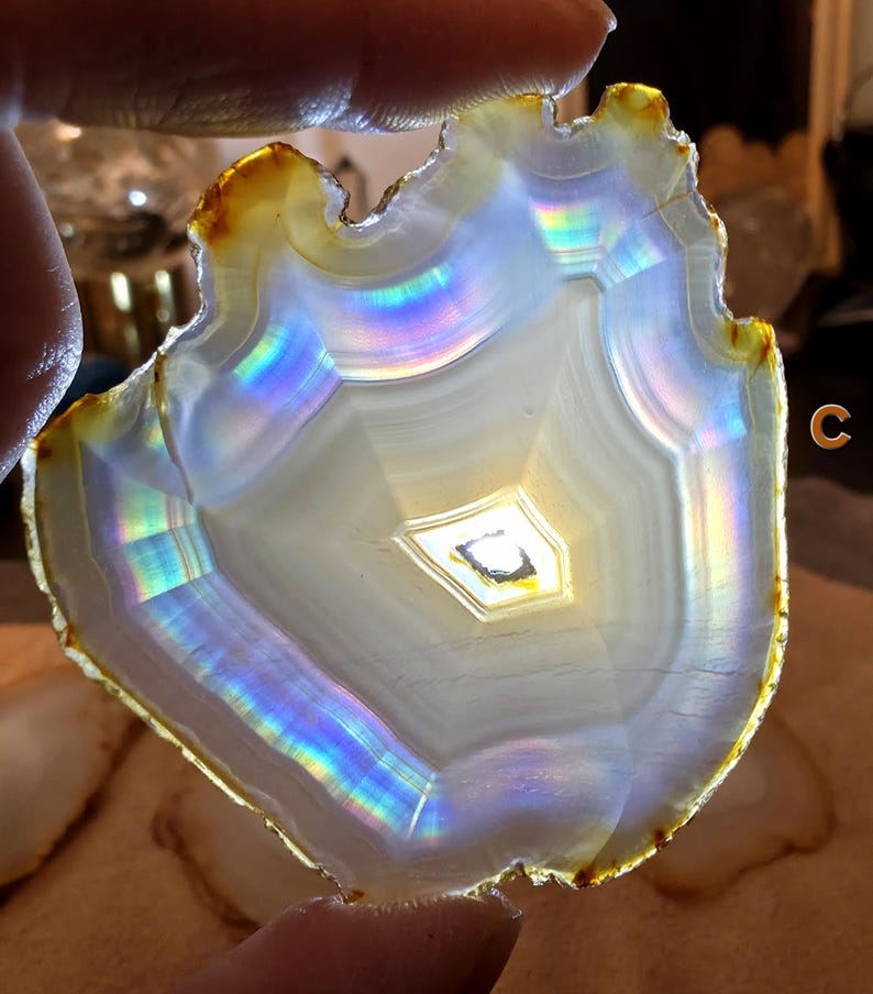 May include: A translucent agate slice with a natural, irregular edge. The stone features concentric bands of white and grey, with iridescent rainbow hues reflecting light. The outer edge has a golden-brown colour. The letter "C" is visible in the image.