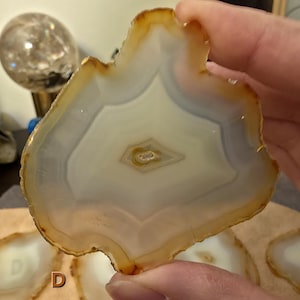 May include: A natural agate slice, showcasing a pattern of white, grey, and amber bands. The polished stone has a central eye-shaped design and a raw, irregular edge. The agate is held in a hand, with other slices in the background, possibly part of a set.