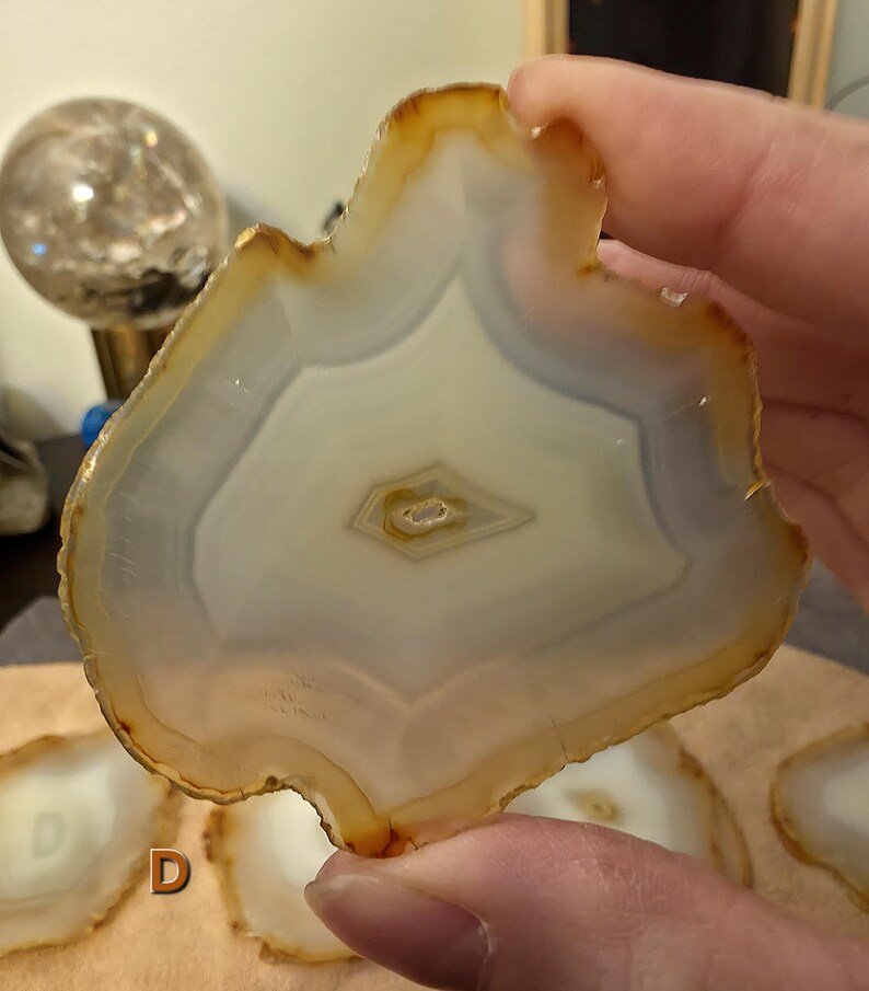May include: A natural agate slice, showcasing a pattern of white, grey, and amber bands. The polished stone has a central eye-shaped design and a raw, irregular edge. The agate is held in a hand, with other slices in the background, possibly part of a set.