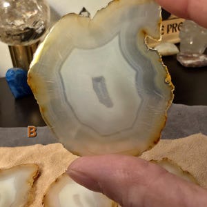 May include: A polished agate slice, displaying concentric bands of white, grey, and tan. The natural stone is held up, revealing its unique patterns and textures. Other agate slices are visible, suggesting a collection of decorative items, with a focus on natural materials.