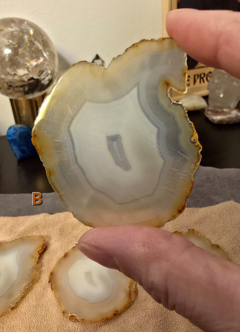 May include: A polished agate slice, displaying concentric bands of white, grey, and tan. The natural stone is held up, revealing its unique patterns and textures. Other agate slices are visible, suggesting a collection of decorative items, with a focus on natural materials.