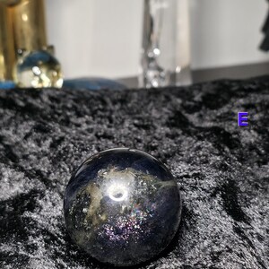 May include: A dark, iridescent sphere with a polished surface. The sphere has a mix of dark blue, purple, and gold colors, with a shimmering, speckled appearance. The sphere is sitting on a black, textured surface.