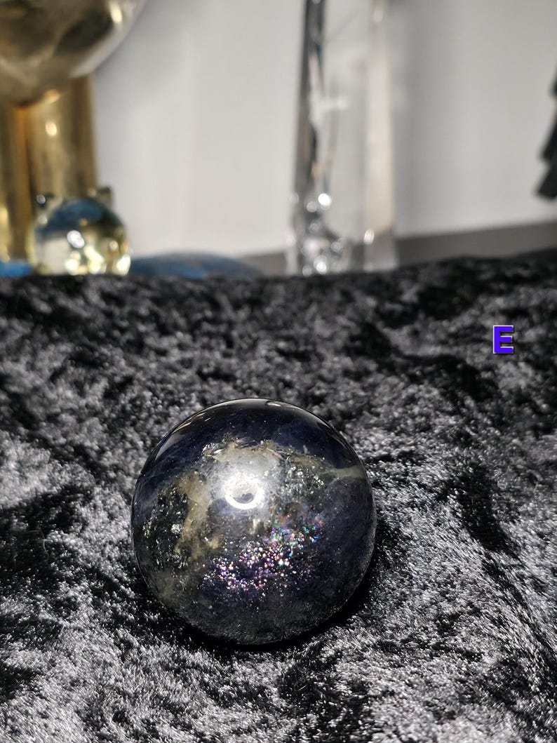 May include: A dark, iridescent sphere with a polished surface. The sphere has a mix of dark blue, purple, and gold colors, with a shimmering, speckled appearance. The sphere is sitting on a black, textured surface.