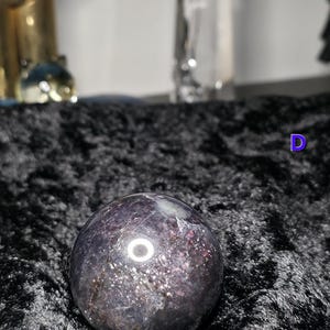 May include: A dark purple crystal sphere with sparkling inclusions. The polished gemstone ball has a reflective surface and sits on a black, textured fabric. The sphere is a decorative item.