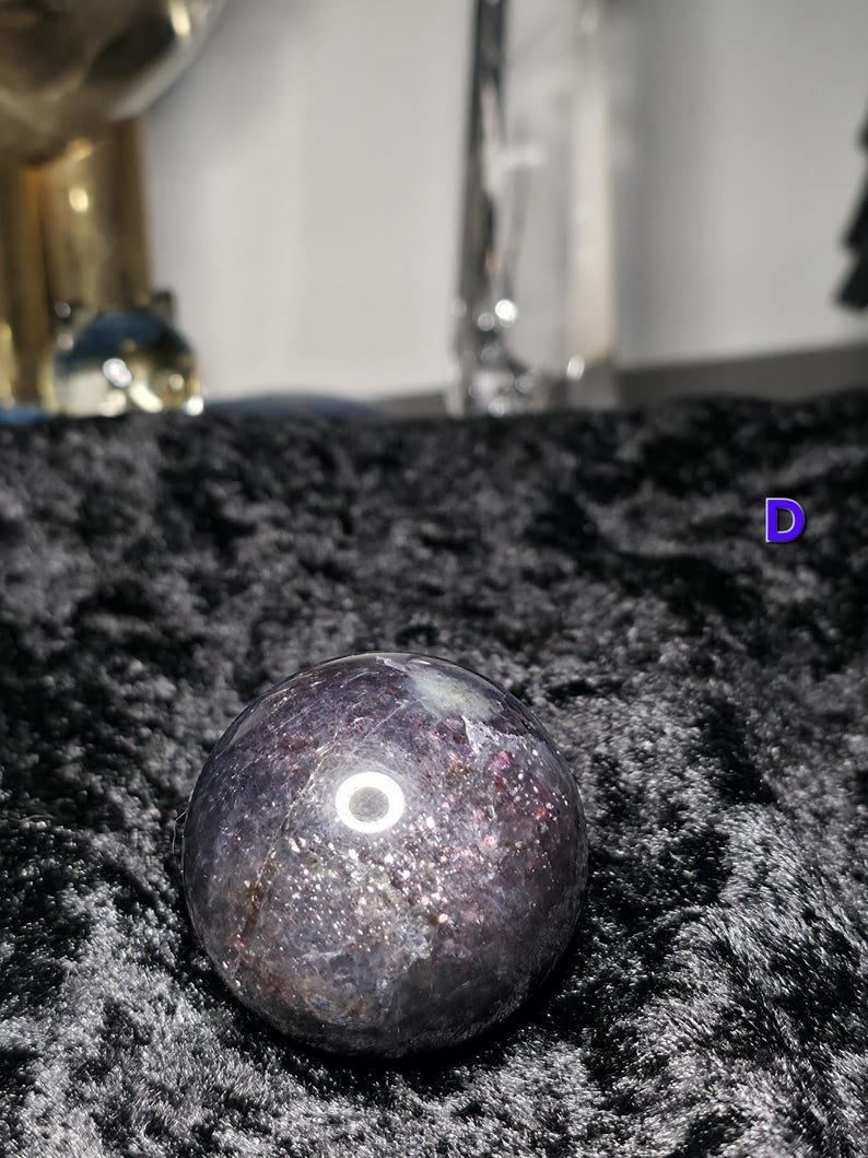 May include: A dark purple crystal sphere with sparkling inclusions. The polished gemstone ball has a reflective surface and sits on a black, textured fabric. The sphere is a decorative item.