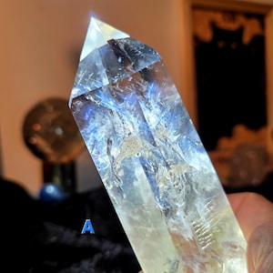 Op de afbeelding: Een heldere kwartskristal met blauwe en gele interne insluitsels. Het kristal wordt in een hand gehouden en wordt verlicht door een helder licht. Het kristal heeft een puntige punt en zichtbare interne structuren. De letter "A" is zichtbaar.