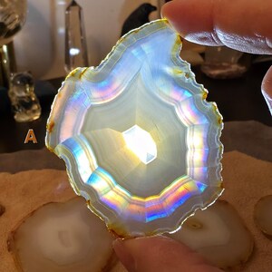 May include: A translucent agate slice with a central, geometric pattern. The stone displays a spectrum of iridescent colours along its edges, including blues, greens, and yellows. The agate is held by a hand, with other agate slices visible in the background.