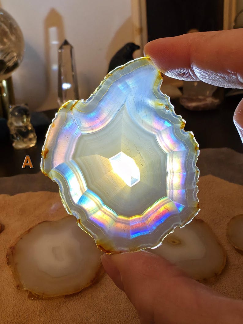 May include: A translucent agate slice with a central, geometric pattern. The stone displays a spectrum of iridescent colours along its edges, including blues, greens, and yellows. The agate is held by a hand, with other agate slices visible in the background.