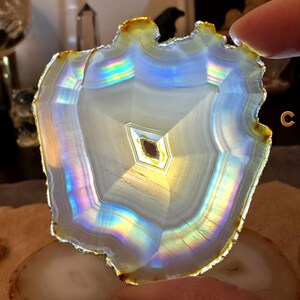May include: A polished agate slice, displaying concentric bands of white, grey, and iridescent colours. The edges have a warm golden hue. The stone is held up, highlighting its natural patterns and textures. The theme is natural stone decor.