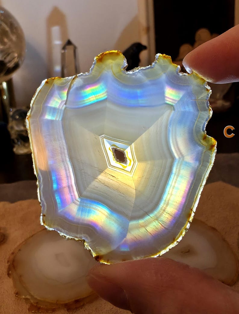 May include: A polished agate slice, displaying concentric bands of white, grey, and iridescent colours. The edges have a warm golden hue. The stone is held up, highlighting its natural patterns and textures. The theme is natural stone decor.